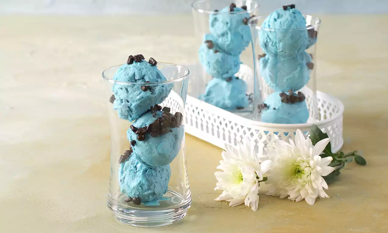 Canlı mavi dondurma topları ince cam bardakta, üzeri çikolata parçaları ile süslenmiş, beyaz bir tepsi ve çiçek detaylarıyla sunulmuş