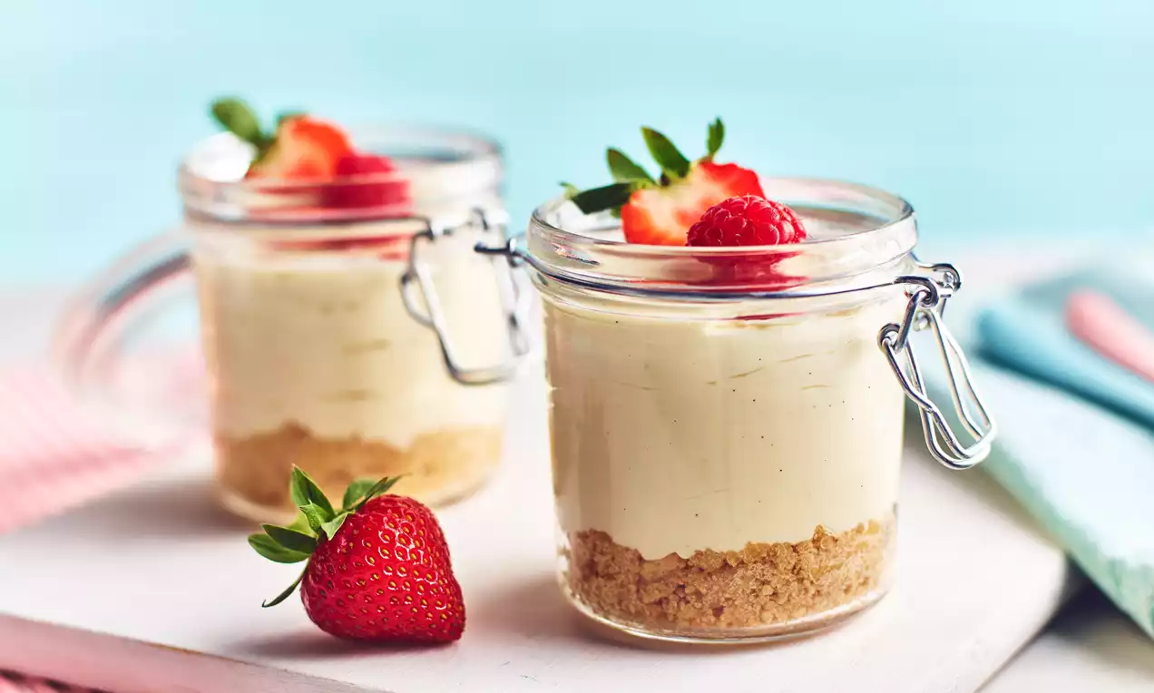Mini vanilla cheesecakes in glass jars with crumbly biscuit bases, creamy pale yellow filling, and fresh strawberries on top