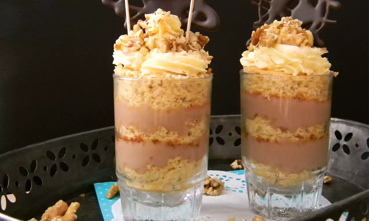 Schichtdessert im Glas mit goldbraunem Biskuit, hellbrauner Creme, Sahnehaube, gehackten Walnüssen und dunkler Schokoladendekoration auf einer Platte
