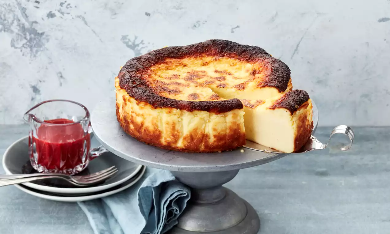 San Sebastian Cheesecake mit goldgelber, karamellisierter Oberfläche und cremiger Füllung auf silberner Tortenplatte, daneben Himbeersauce