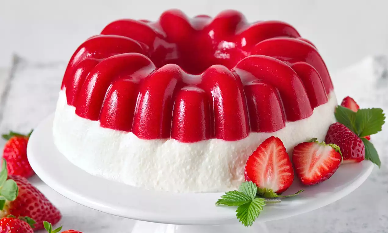 Lesklá červená bábovka z jahodového želé na nadýchanej bielej pene, ozdobená čerstvými jahodami a zelenými lístkami na bielom podnose