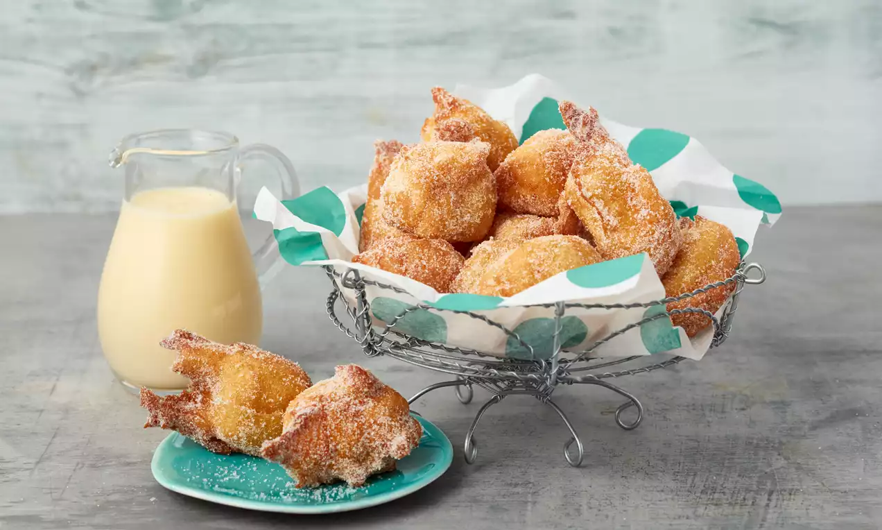 Goldbraune, gezuckerte Faschingskrapfen in einem silbernen Drahtkorb mit gemustertem Papier, daneben eine Karaffe mit heller Weinsoße