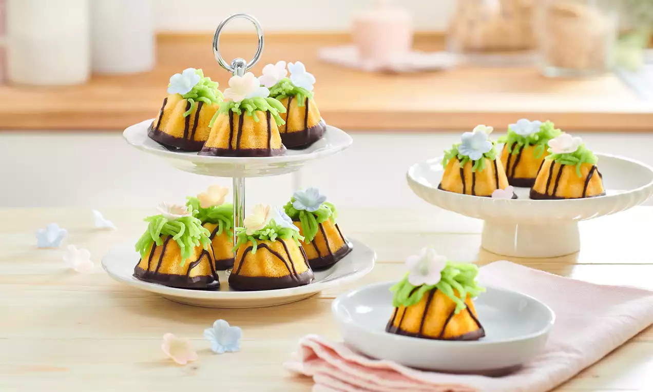 Kleine goldgelbe Gugelhupfe mit grüner Creme und pastellfarbenen Blüten verziert, auf Etageren und Tellern angerichtet