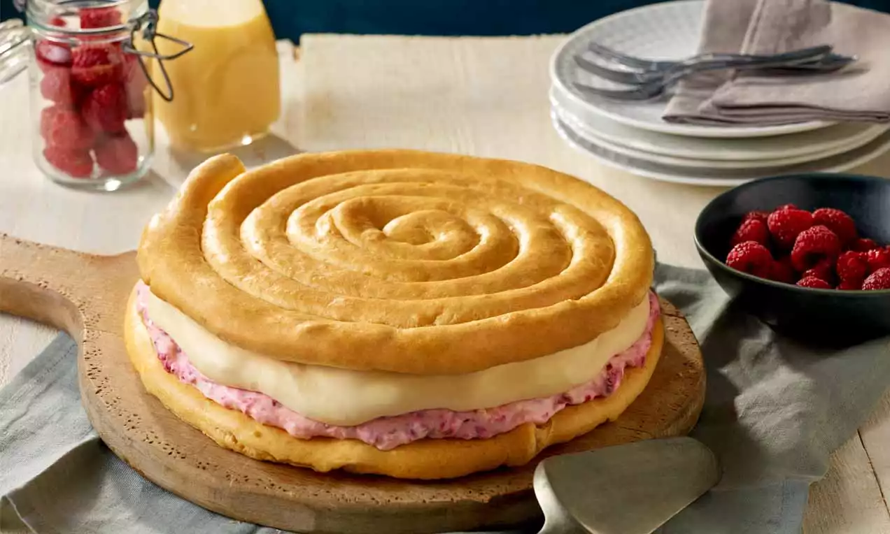 Brandteig-Torte in spiralförmiger Optik mit heller Sahnecreme und rosa Himbeerfüllung auf einem Holzbrett, umgeben von frischen Himbeeren