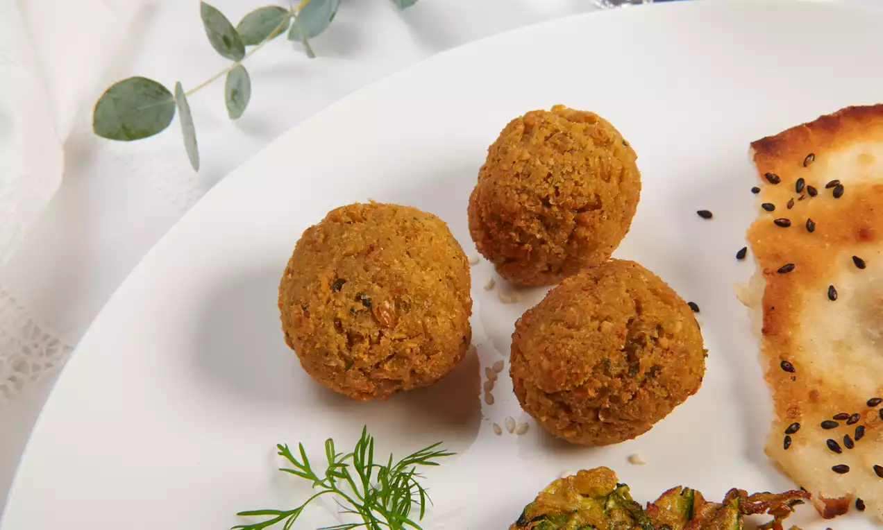 Drei goldbraune, kugelförmige Falafel mit leicht krümeliger Textur, angerichtet neben Fladenbrot und frischen Kräutern auf einem weißen Teller