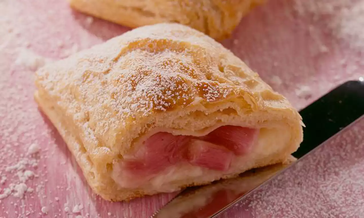 Knusprige, goldene Teigtasche mit zartem Puderzucker bestäubt, gefüllt mit cremiger Vanille und rosa Rhabarberstücken auf rosa Untergrund