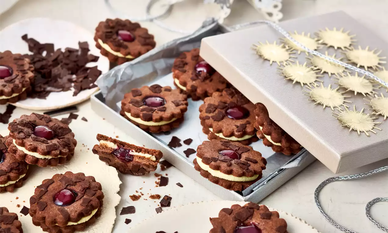 Dunkelbraune Doppeldecker-Kekse mit gezacktem Rand, heller Cremefüllung und rotem Kirschglanz in der Mitte, umgeben von Schokoladensplittern