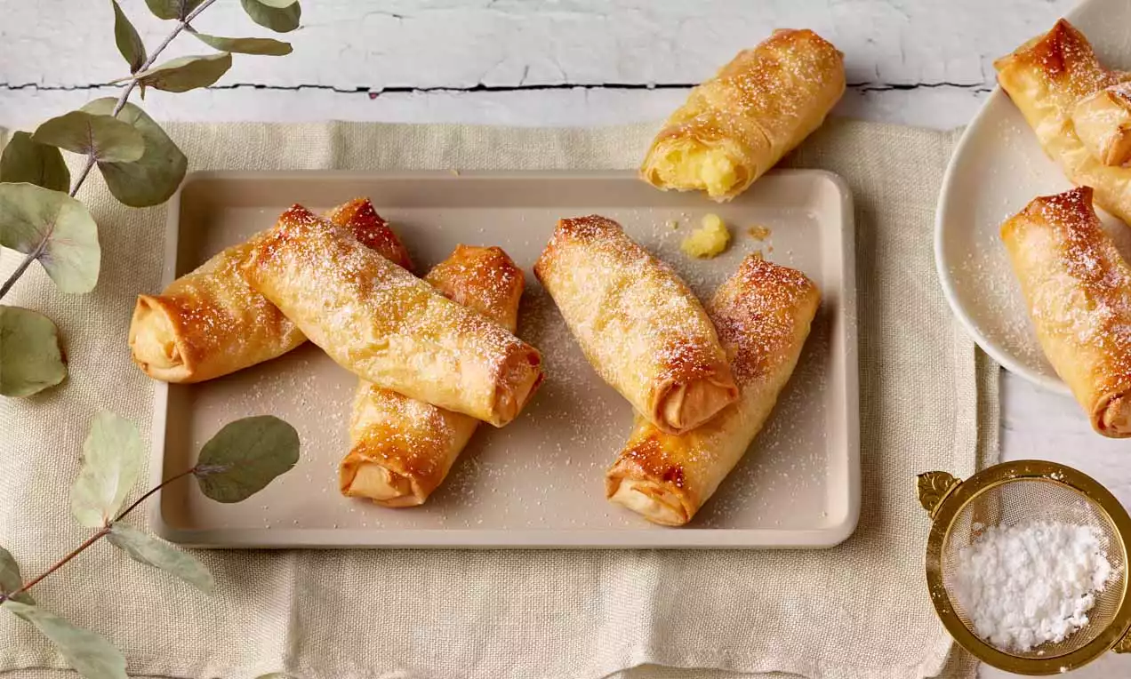 Goldbraune Puddingröllchen aus knusprigem Teig, teils aufgebrochen mit sichtbarer Füllung, bestäubt mit Puderzucker auf einem beigen Tablett