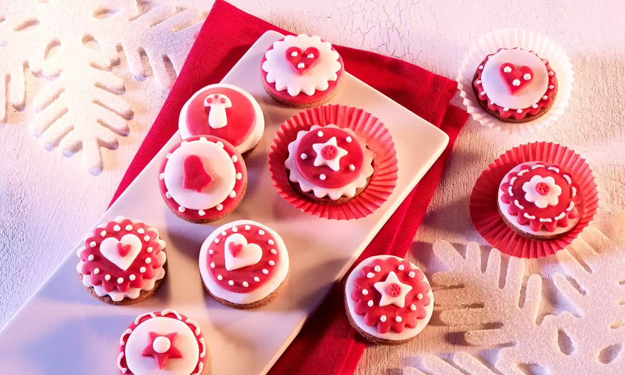Biscotti decorati con glassa rossa e bianca a tema natalizio, con motivi di cuori e stelle, su un piatto bianco e tovagliolo rosso