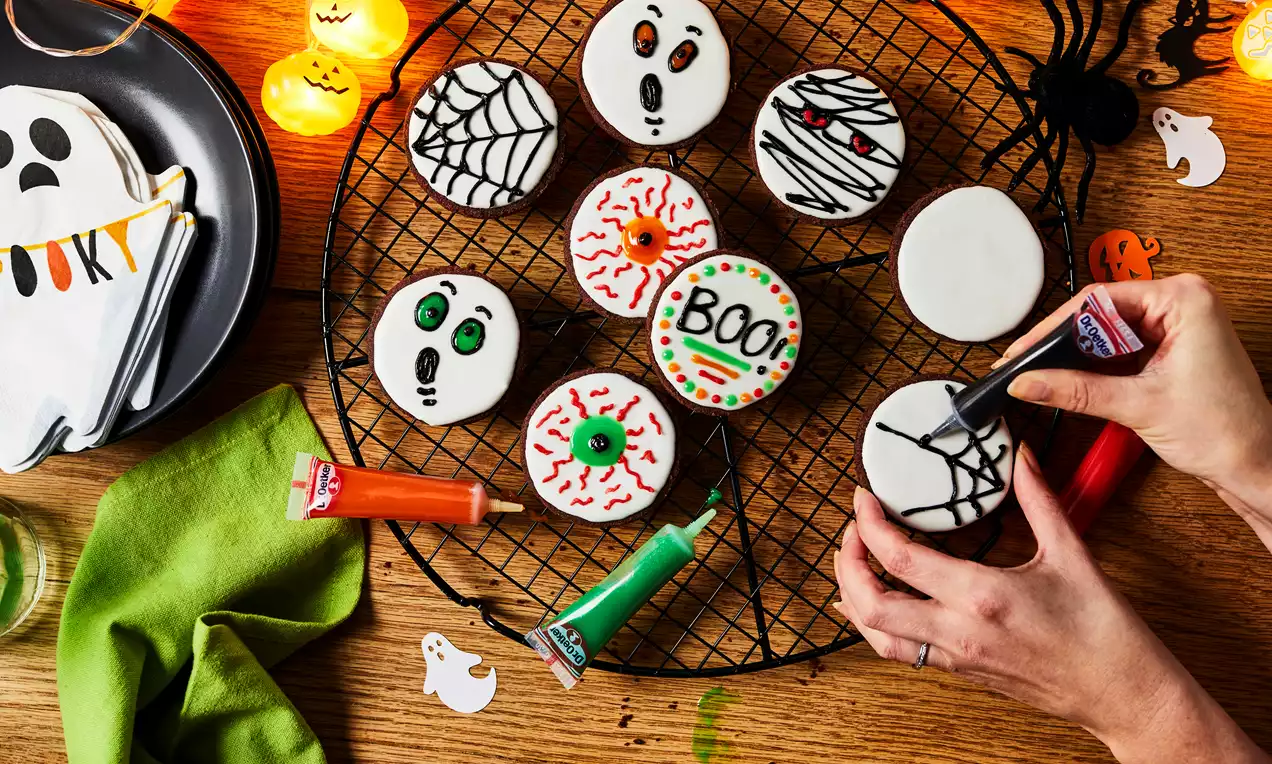 Round Halloween biscuits with white icing, decorated with colourful spooky designs, arranged on a black wire rack with ghost cut-outs nearby