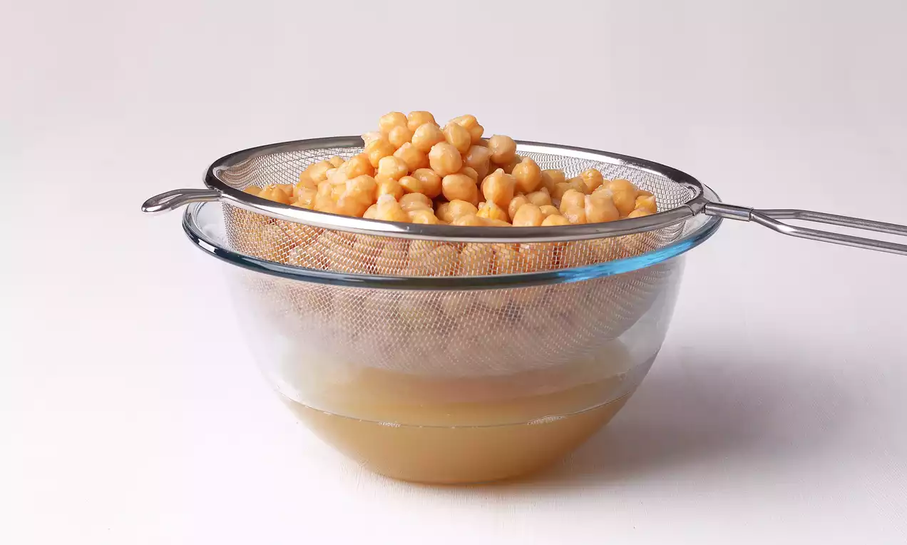 Paslanmaz metal süzgeçte süzülen altın rengi nohutlar, altındaki geniş cam kasede biriken açık sarı renkli nohut suyu