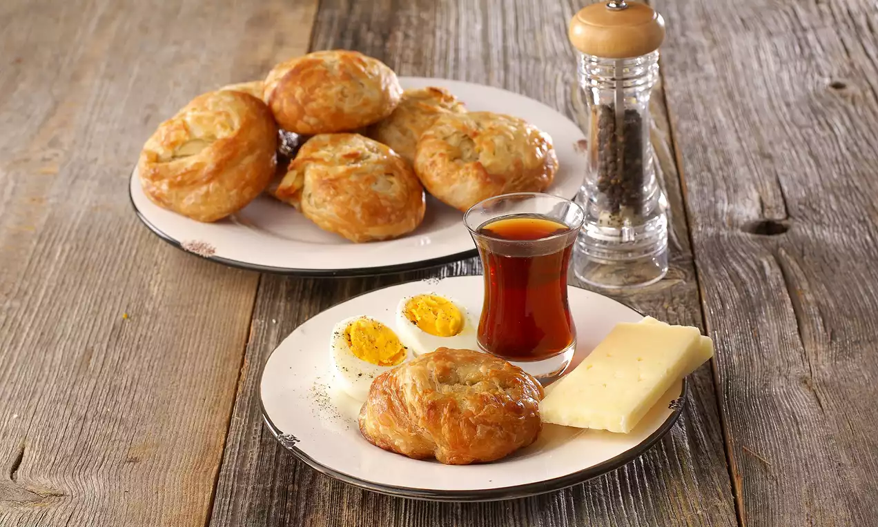 Altın rengi kabarık boyozlar ahşap zemin üzerinde, yanında dilimlenmiş peynir, haşlanmış yumurta ve ince belli bardakta çay ile sunulmuş