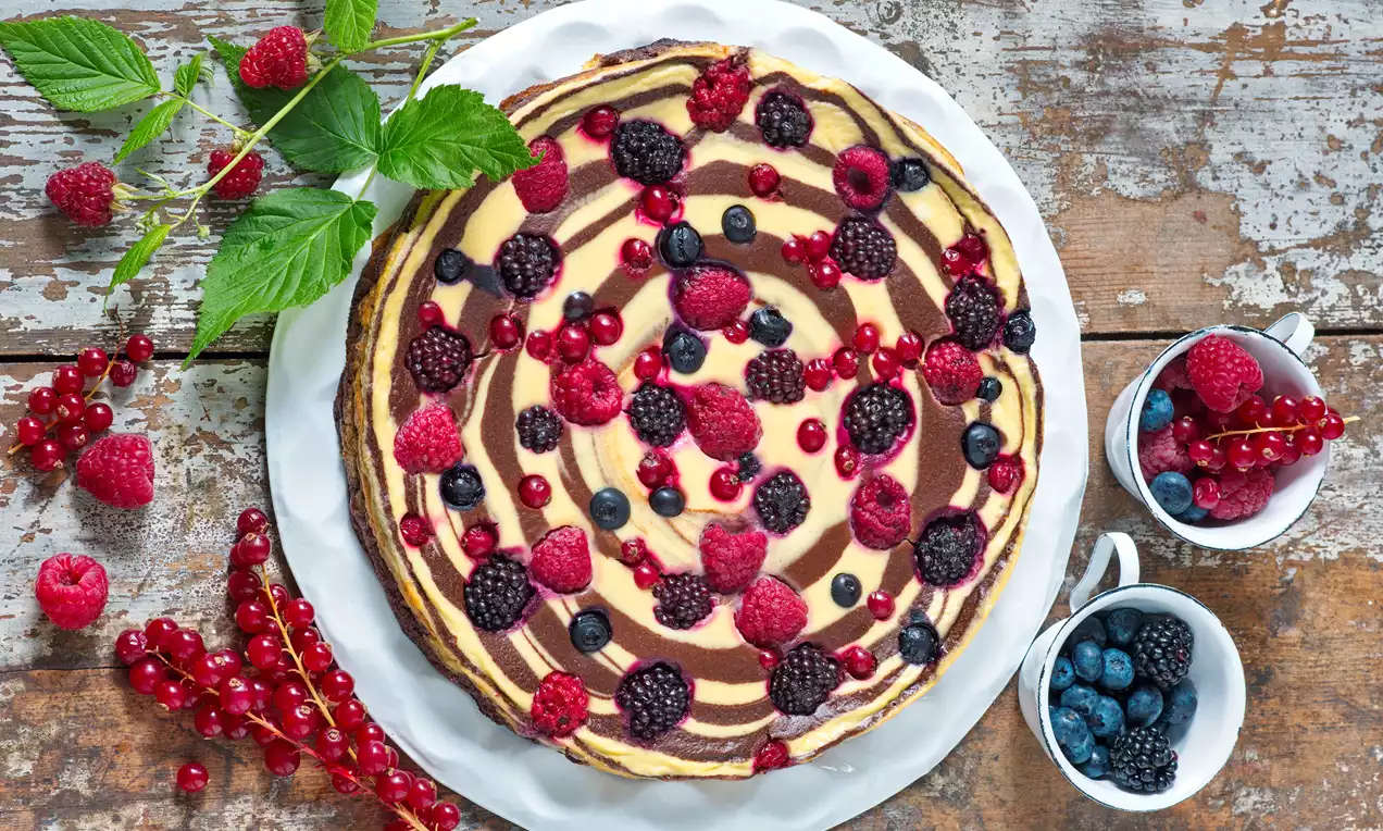 Dvoubarevný tvarohový dort se spirálovým vzorem v tónech krémové a kakaové, ozdobený malinami, ostružinami, rybízem a borůvkami na bílém talíři