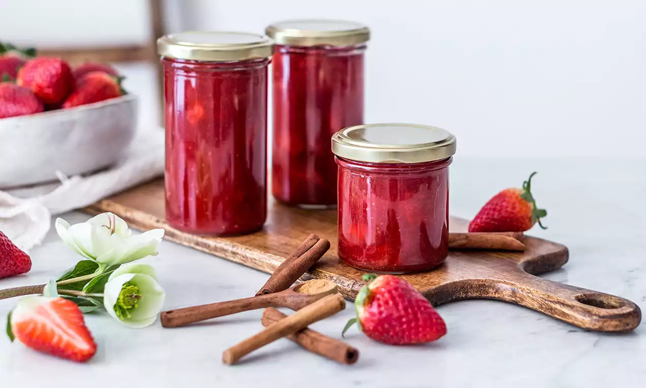 Tri sklenené poháre s lesklým červeným džemom na drevenom podnose, obklopené čerstvými jahodami, škoricovými paličkami a bielymi kvetmi