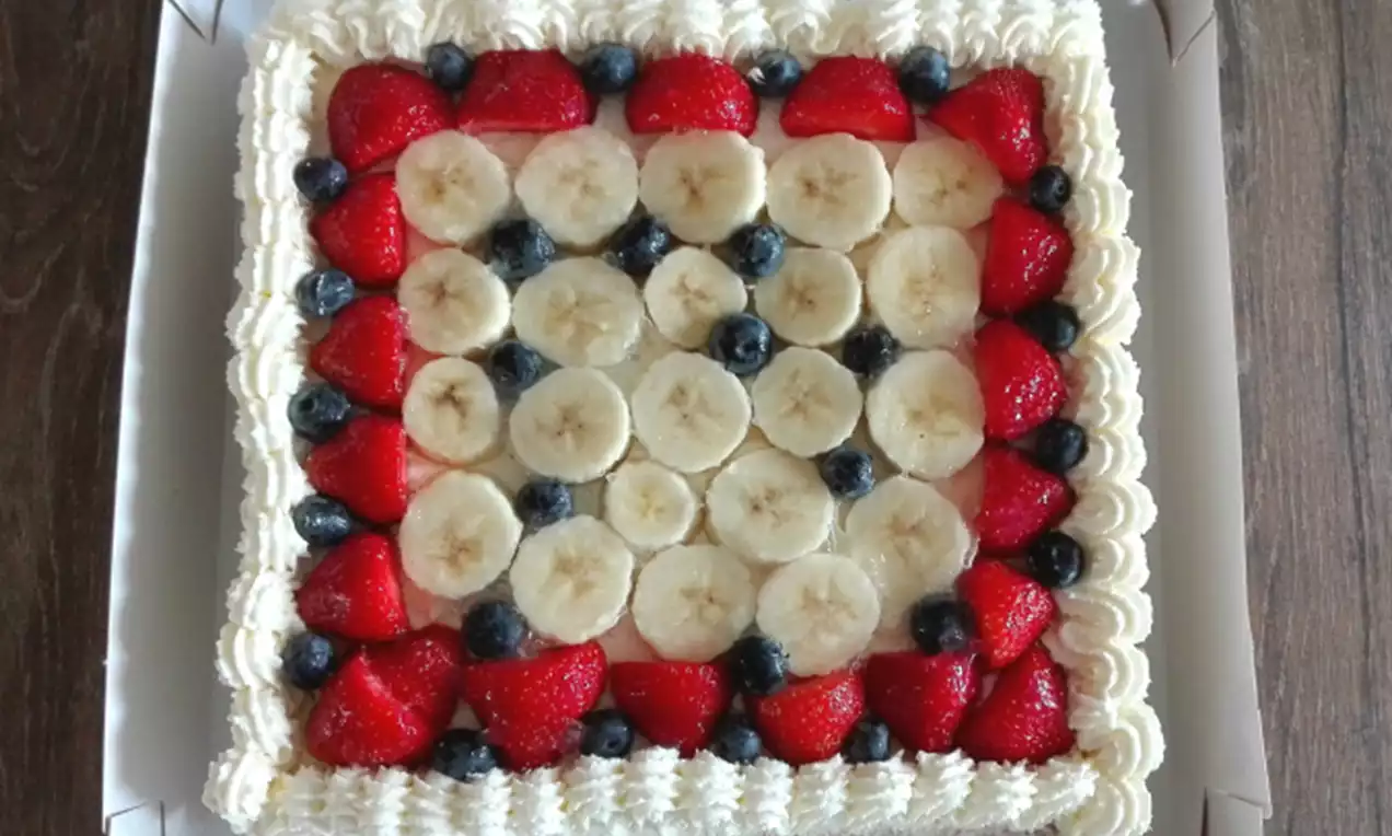 Vierkante taart met witte slagroomranden, bedekt met lagen plakjes banaan, sappige aardbeien en blauwe bessen in een kleurrijk patroon