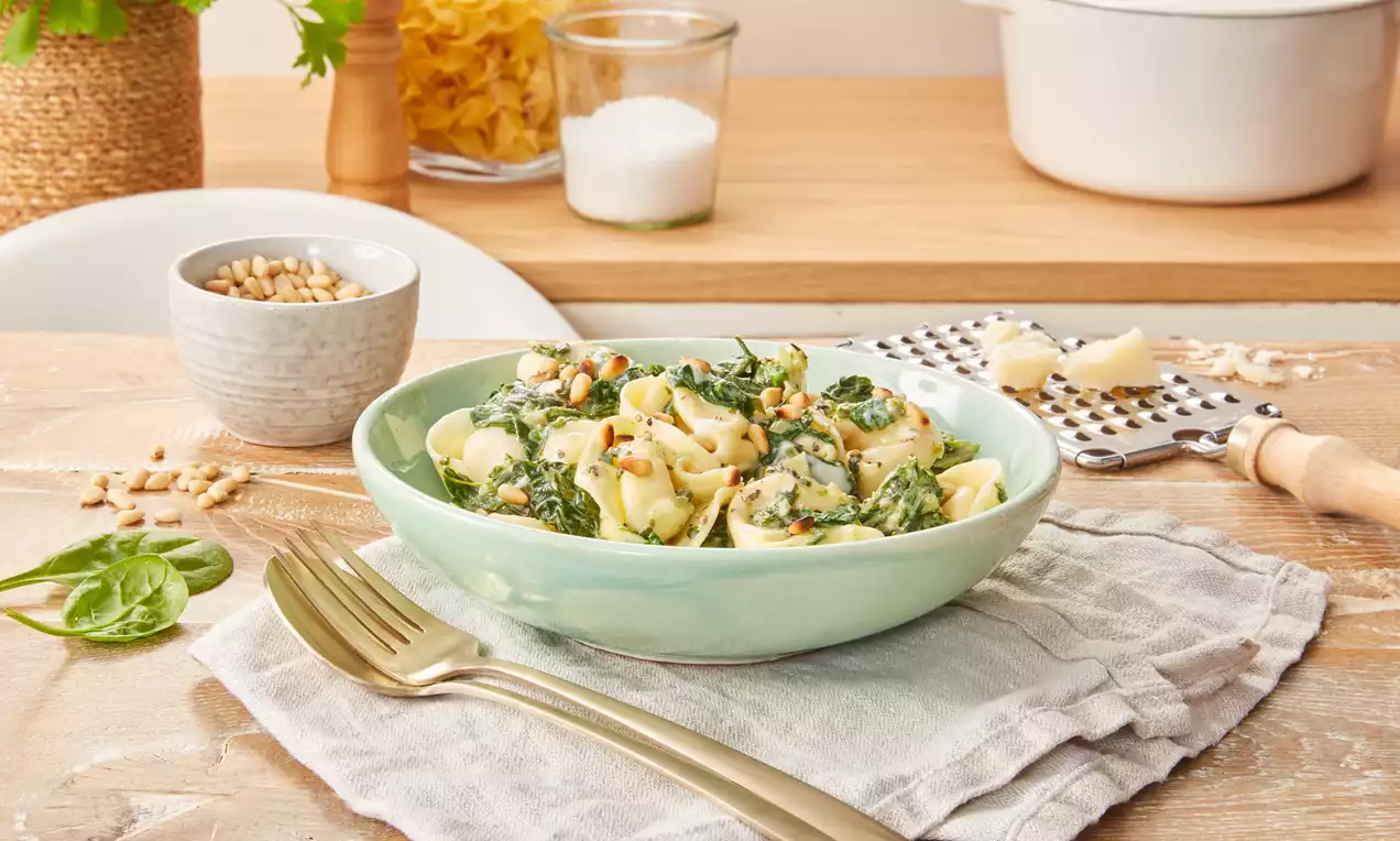 Tortelloni mit hellgelber Spinat-Käsesoße und goldbraunen Pinienkernen in einer pastellgrünen Schale auf grauem Tuch drapiert
