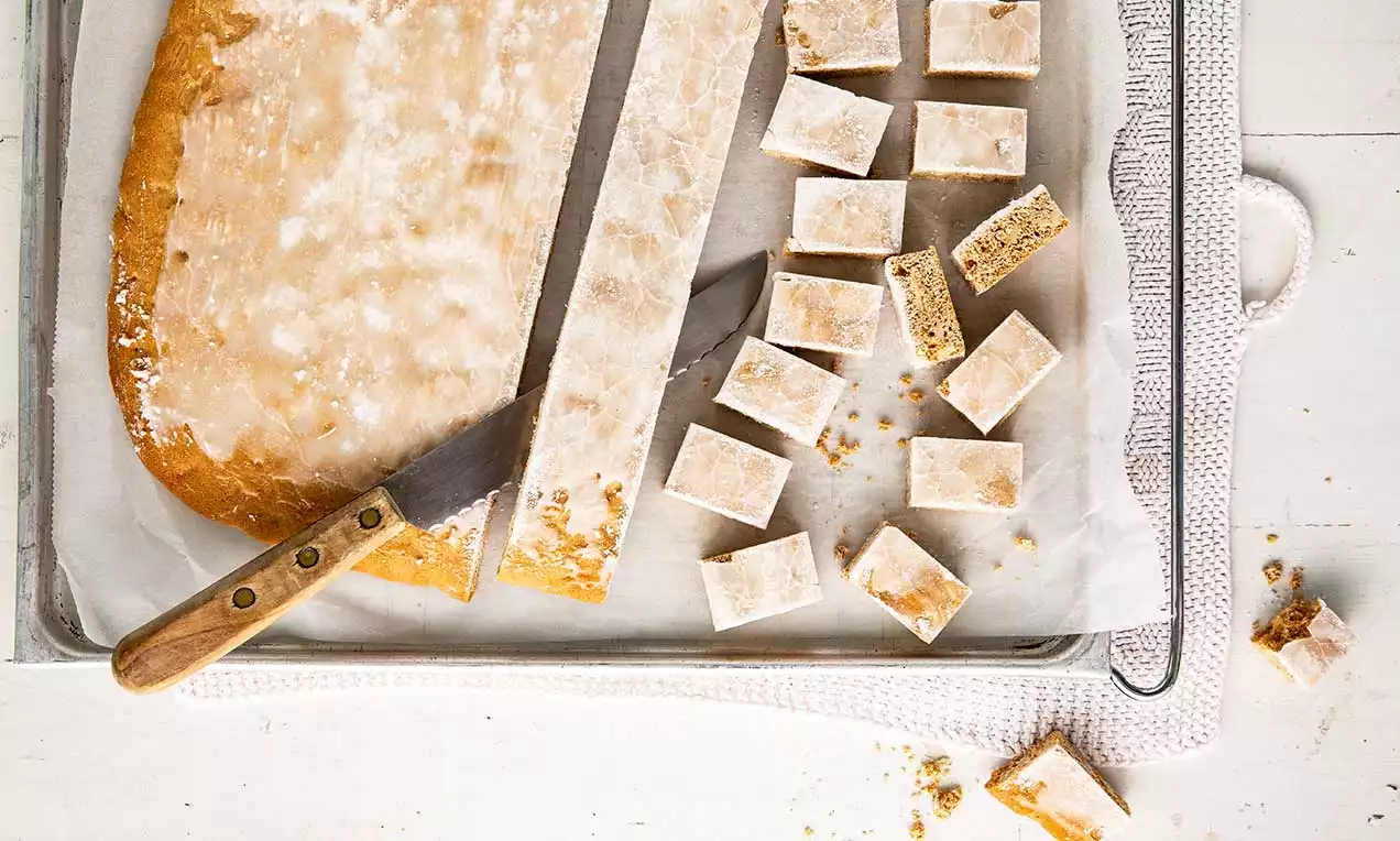 Cubetti di biscotto rettangolari color caramello ricoperti da una glassa sottile bianca, disposti su carta da forno accanto a un coltello