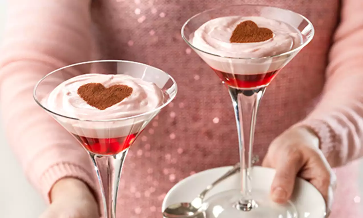 Copas de cristal con capas de gelatina roja, crema rosada suave coronada con cacao en forma de corazón, sostenidas sobre fondo brillante