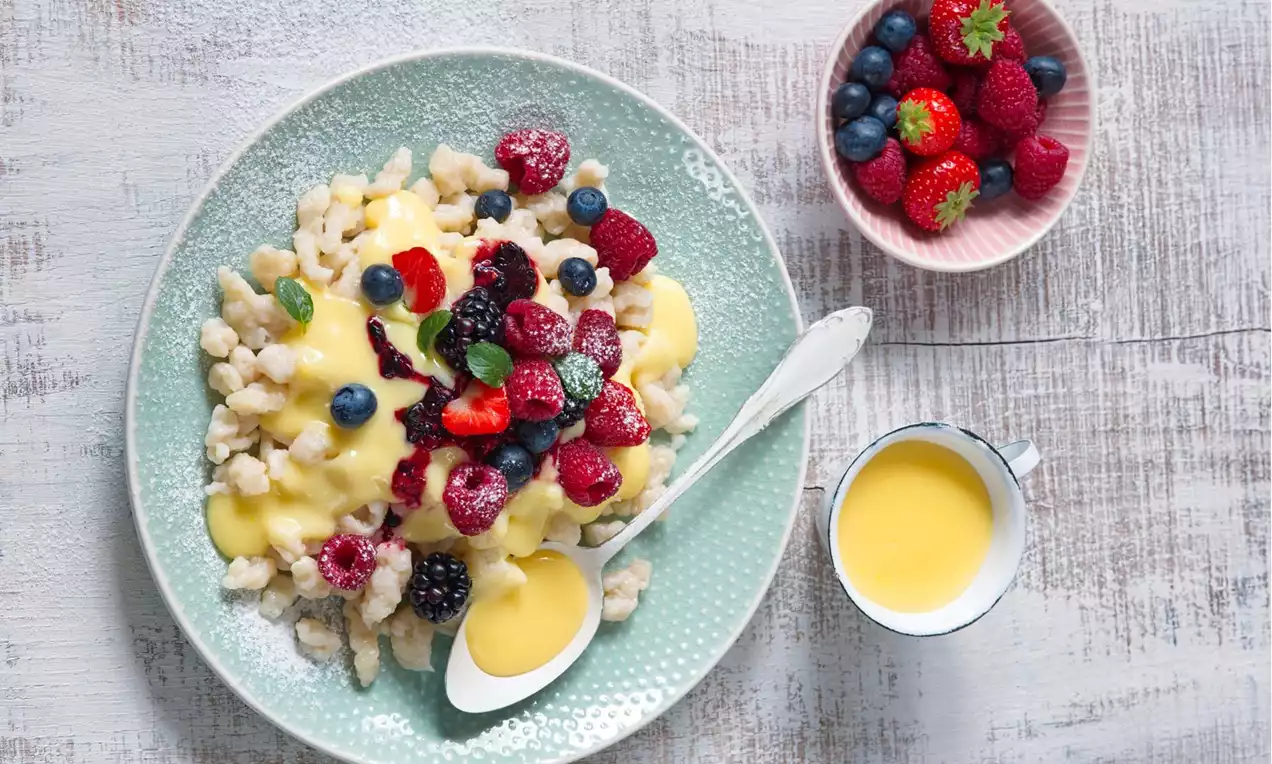 Halušky svetlej farby na zelenkastom tanieri, poliate jemným žltým krémom, ozdobené čerstvým červeným a modrým ovocím s lístkami mäty
