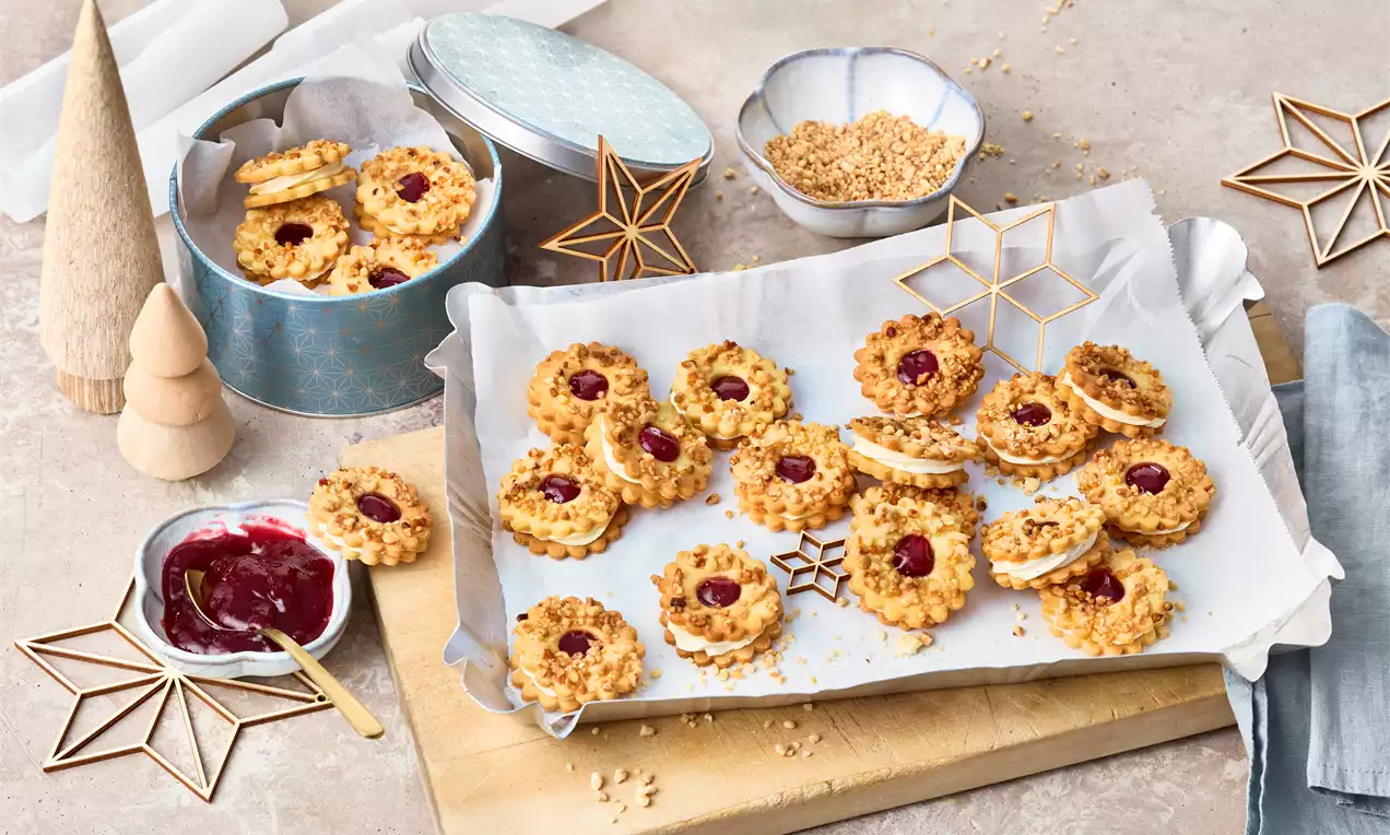 Runde Frankfurter-Kranz-Plätzchen mit goldbraunem Knusperrand, roter Gelee-Füllung und Krokant auf Backpapier mit dekorativen Sternen