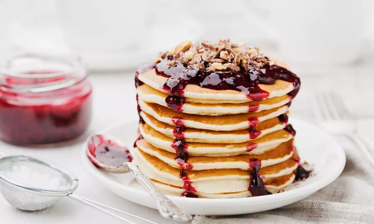 Golden pancakes stacked high with glossy raspberry jam dripping down, topped with crunchy granola on a white plate