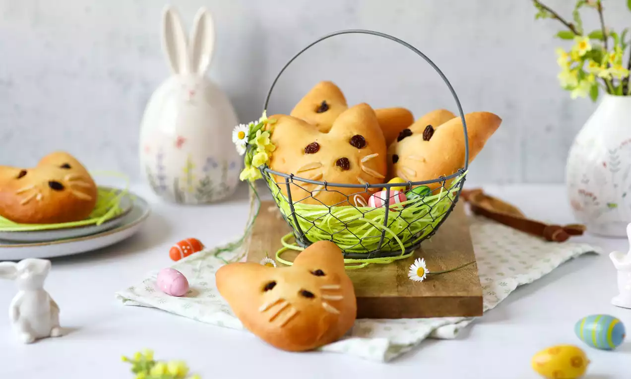 Goldbraune Hasenkopfform-Brotstücke mit glänzenden Rosinenaugen in einem Drahtkorb mit grünem Gras und kleinen Blumen dekoriert