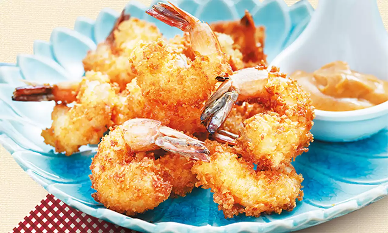 Golden crispy breaded shrimp with curled tails arranged on a blue plate, served with a creamy dipping sauce in a white bowl