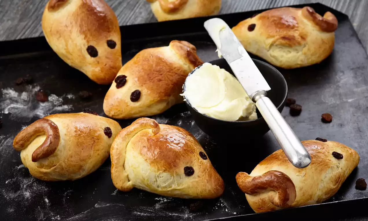 Goldbraune Hefemäuse mit rosinenartigen Augen und geschwungenen Schwänzen auf dunklem Blech neben einer Schale mit cremiger Butter