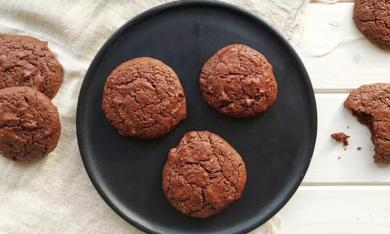 Guľaté kakaové sušienky s popraskaným povrchom, jemne lesklé, rozložené na čiernom tanieri, s kúskami pekanových orechov a drobkami na bielej látke.
