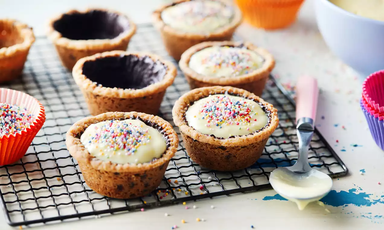 Cookie cups met bruine koekbodems gevuld met romige witte vulling, bestrooid met kleurrijke hagelslag op een metalen koelrek