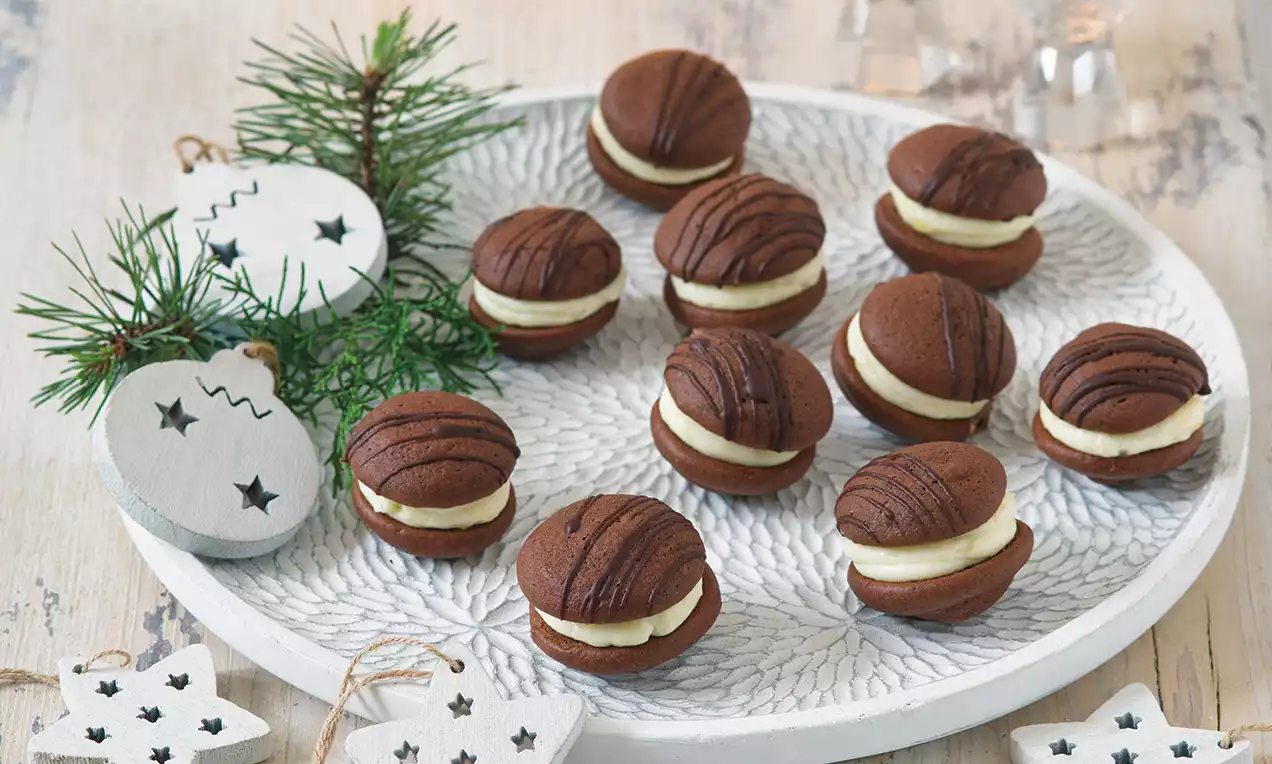 Čokoládové kulaté sušenky s jemným krémem uvnitř, zdobené tmavými čáry, uspořádané na bílém vzorovaném talíři s větvičkou jehličnanu
