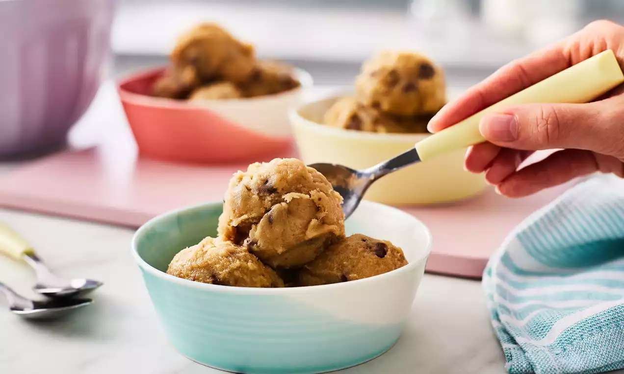 Scooped mounds of golden-brown cookie dough with chocolate chips in a pastel blue bowl, surrounded by bowls and a striped napkin