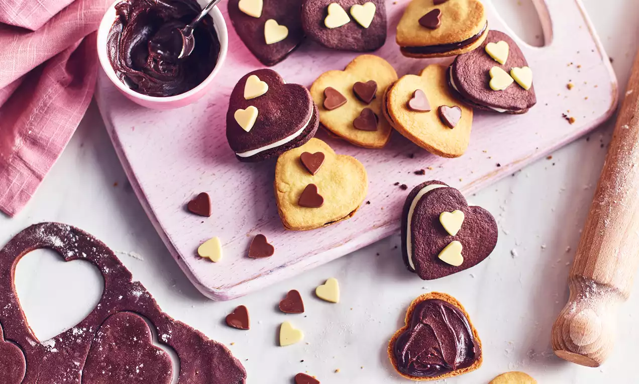 Heart-shaped chocolate and vanilla shortbread sandwiches filled with ganache, adorned with small white and brown heart decorations