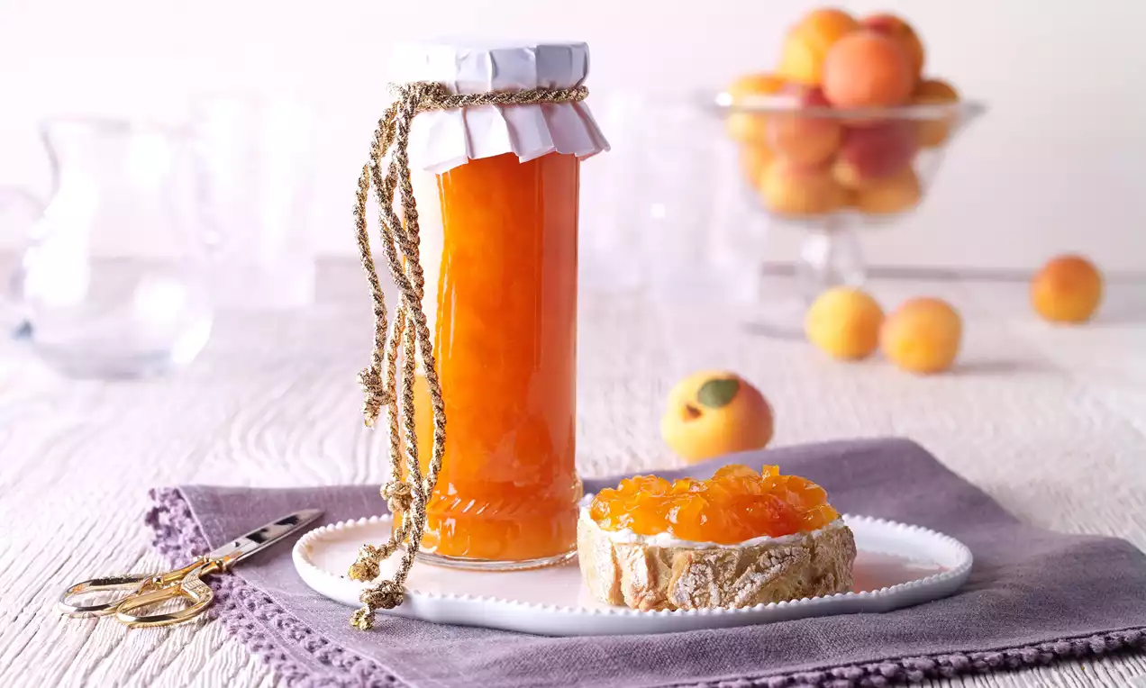 Dulceață de caise într-un borcan înalt decorat cu sfoară, alături de un miez de pâine uns cu gem pe un șervet mov și caise proaspete în fundal