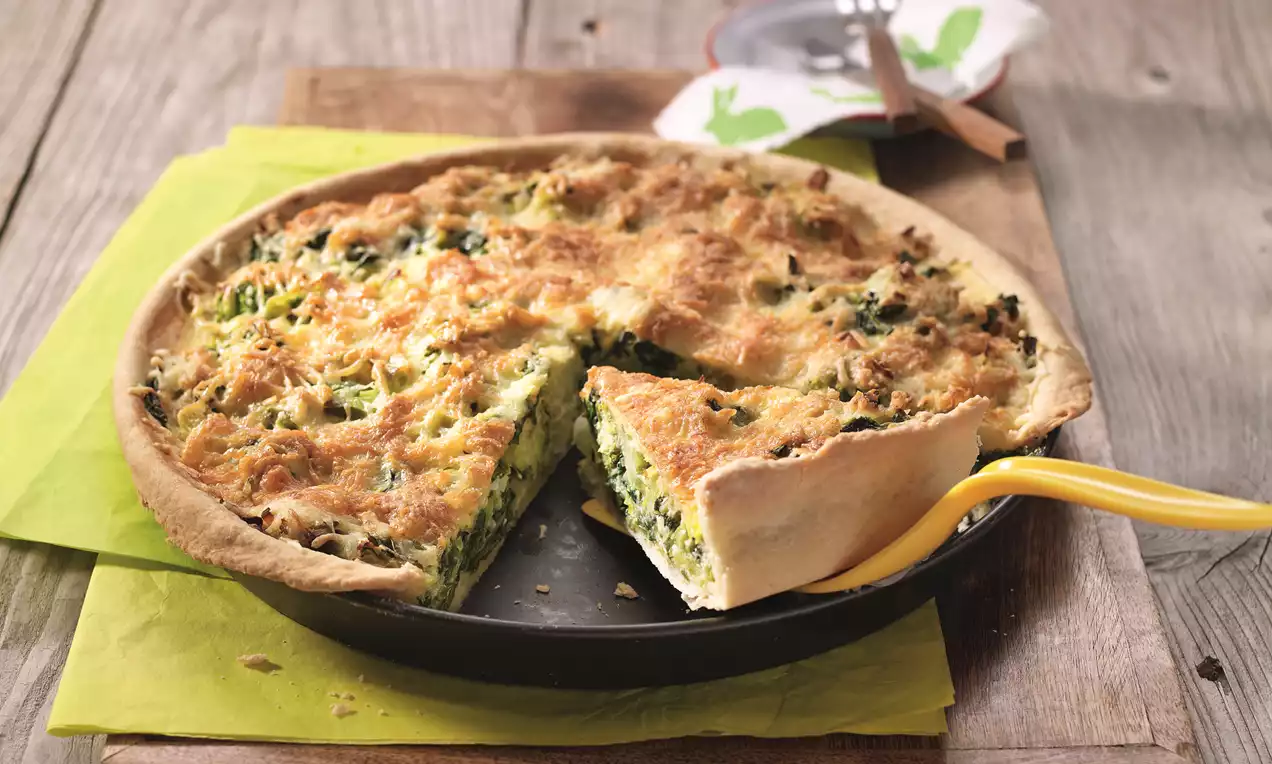 Goldbraune Wähe mit grüner Füllung aus Gemüse und Käse in schwarzer Backform, angeschnitten, auf einem Holztisch mit gelber Serviette drapiert