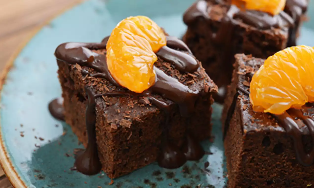 Square-shaped, rich brown chocolate brownies drizzled with dark chocolate sauce and topped with glossy orange mandarin segments on a teal plate