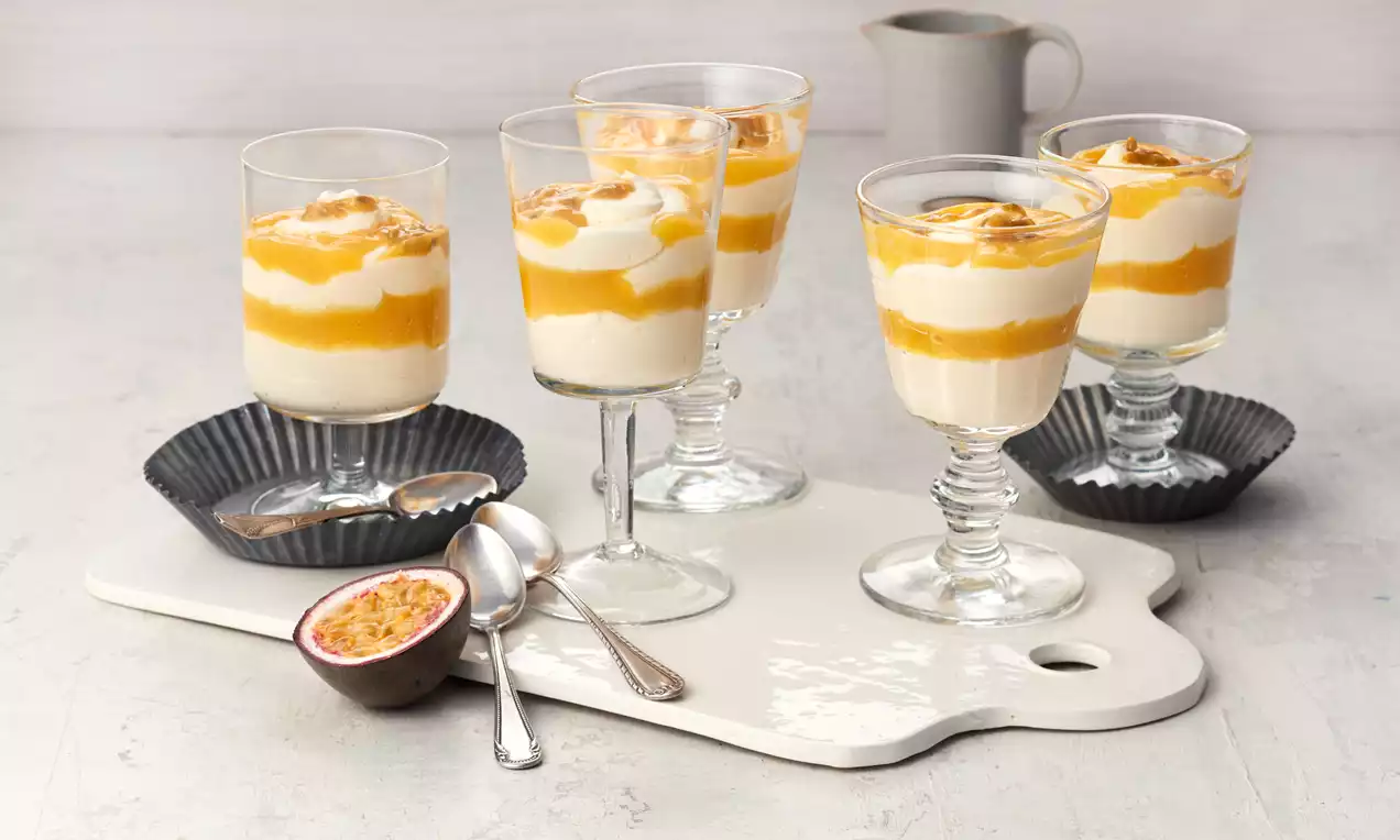 Schichtdessert aus gelber Fruchtsoße und weißer Creme in hohen Gläsern, garniert mit Karamellbröseln, auf heller Servierplatte angerichtet