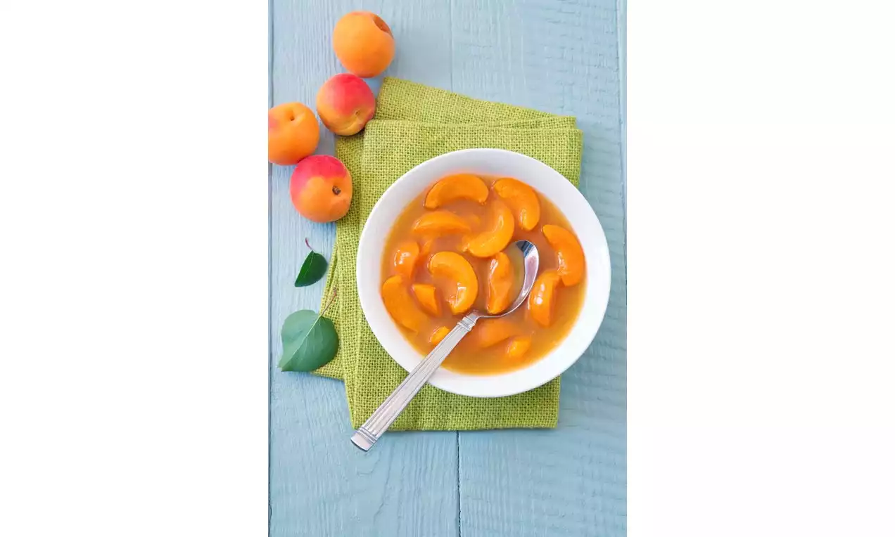 Studená meruňková polievka v bielej miske s oranžovými plátkami meruniek, položená na zelenom obrúsku na svetlom drevenom pozadí