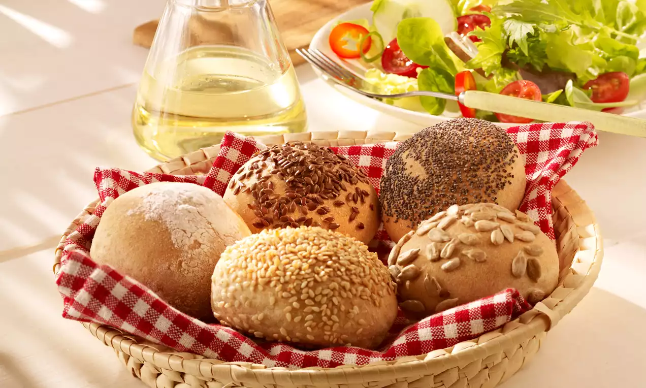 Petits pains aux graines dorés, croûte saupoudrée, disposés sur un tissu à carreaux rouges dans un panier tressé avec une salade fraîche