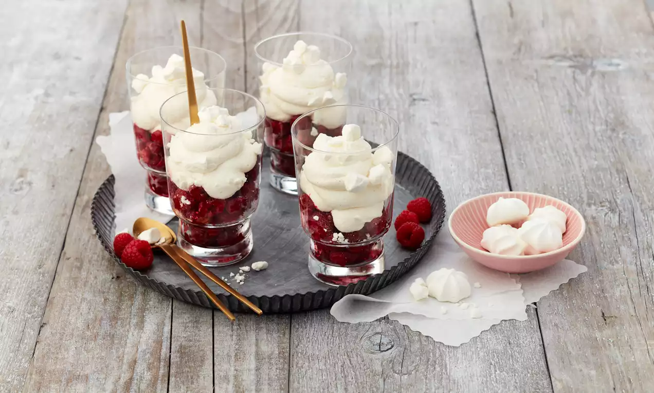 Gläser mit weißer Sahnehaube auf roten Himbeeren, dekoriert mit kleinen Baiserstücken, auf grauem Teller mit Himbeeren und goldenen Löffeln