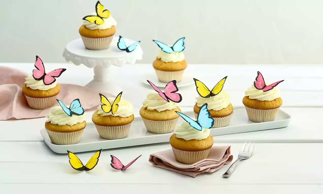 Muffini zlatnožute boje s bijelom kremom i ukrasima u obliku šarenih leptira, raspoređeni na pladnjevima i stalak na bijelom stolu
