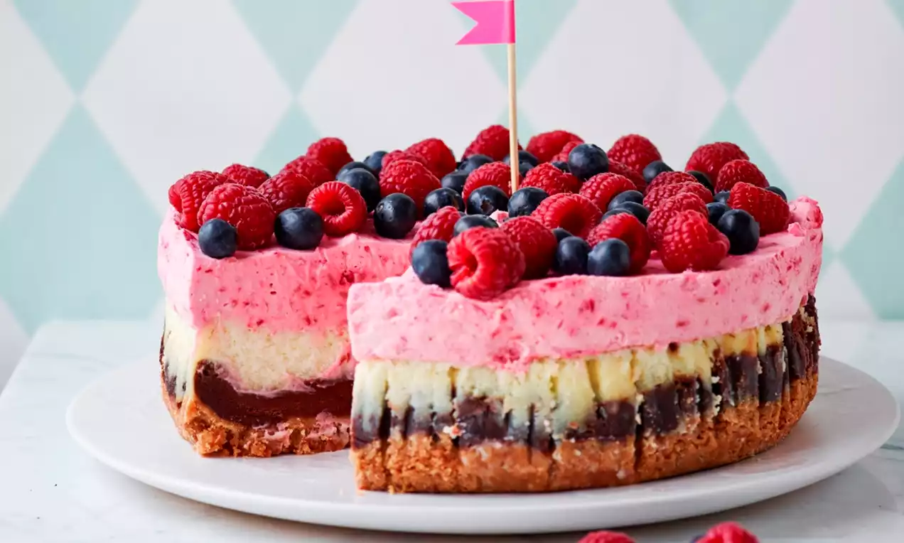 Värikäs juustokakku vaaleanpunaisella marjakerroksella, punaisten vadelmien ja tummansinisten mustikoiden täplittämänä valkoisella lautasella
