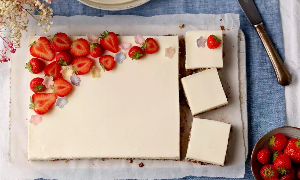 Rechteckige Holunderblütentorte mit weißer Creme, Dekor aus roten Erdbeeren und bunten Blüten auf hellem Backpapier mit Messer und Schale