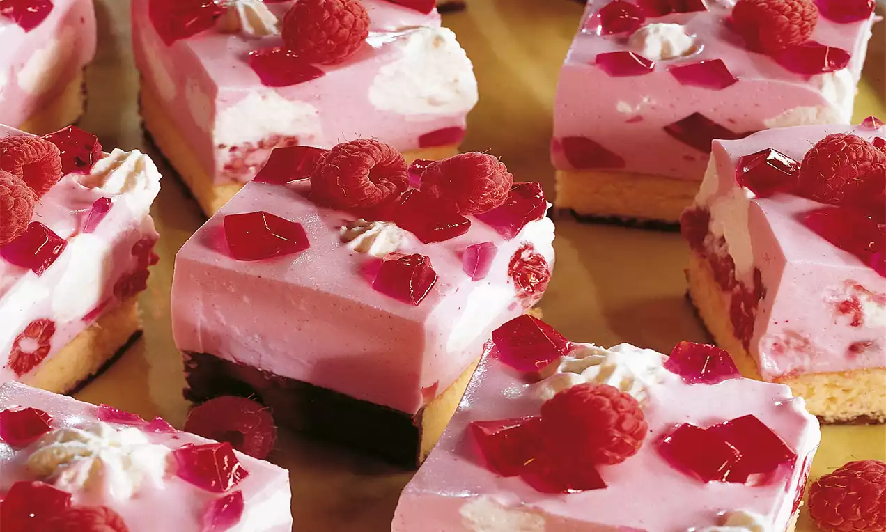 Würfel mit rosafarbener Creme und rotem Götterspeise-Gelee auf hellem Biskuitboden, garniert mit frischen Himbeeren und Sahnetupfern