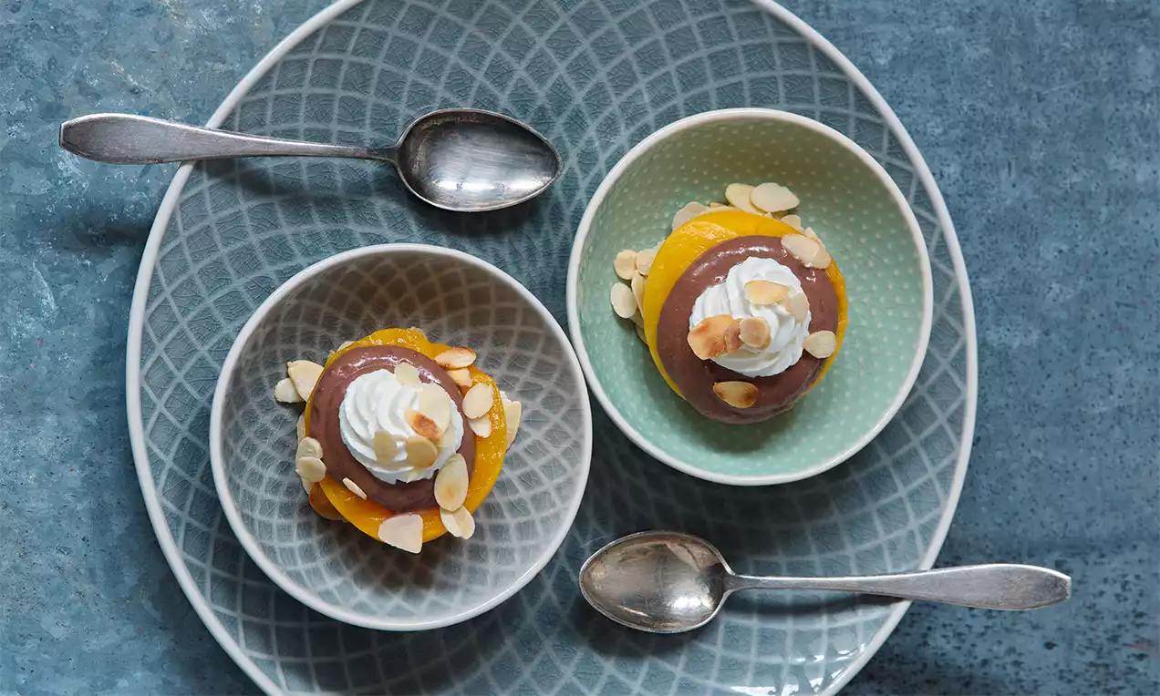 Čokoládový krém na plátkoch šťavnatej oranžovej broskyne, ozdobený bielou šľahačkou a mandľovými plátkami v miskách na modrom tanieri
