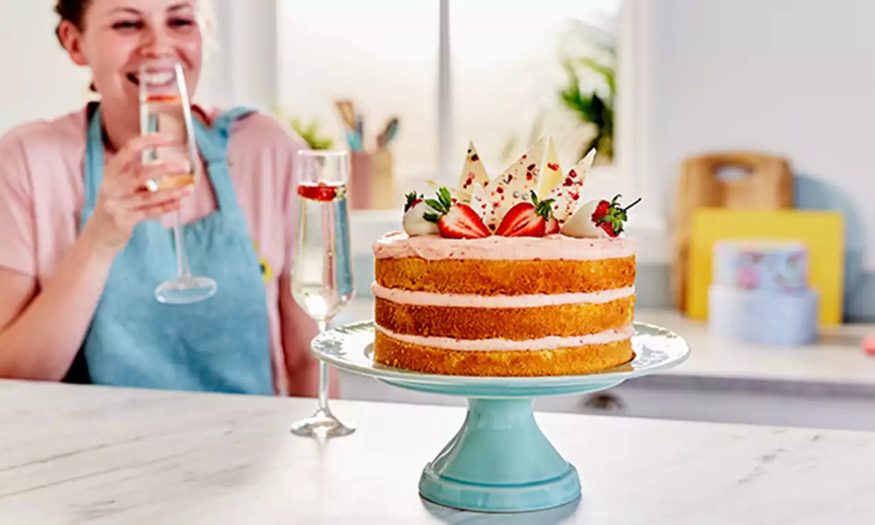 Three-layered golden sponge cake with pale pink frosting, topped with sliced strawberries, edible petals, and white chocolate shards on a light blue cake stand
