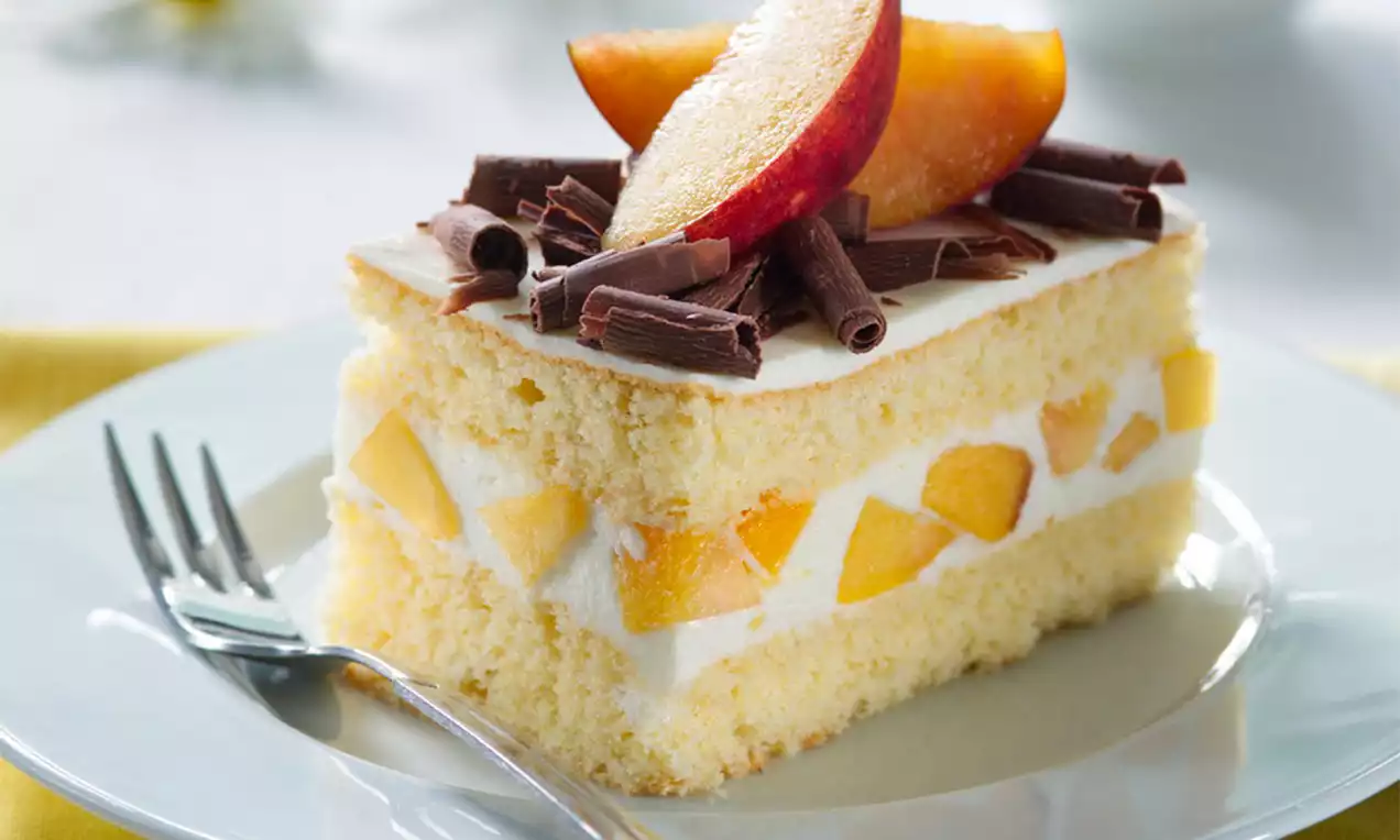 Nadýchaný světlý piškot s vrstvou tvarohového krému a šťavnatých broskví, ozdobený plátky broskví a tmavou čokoládou na bílém talíři