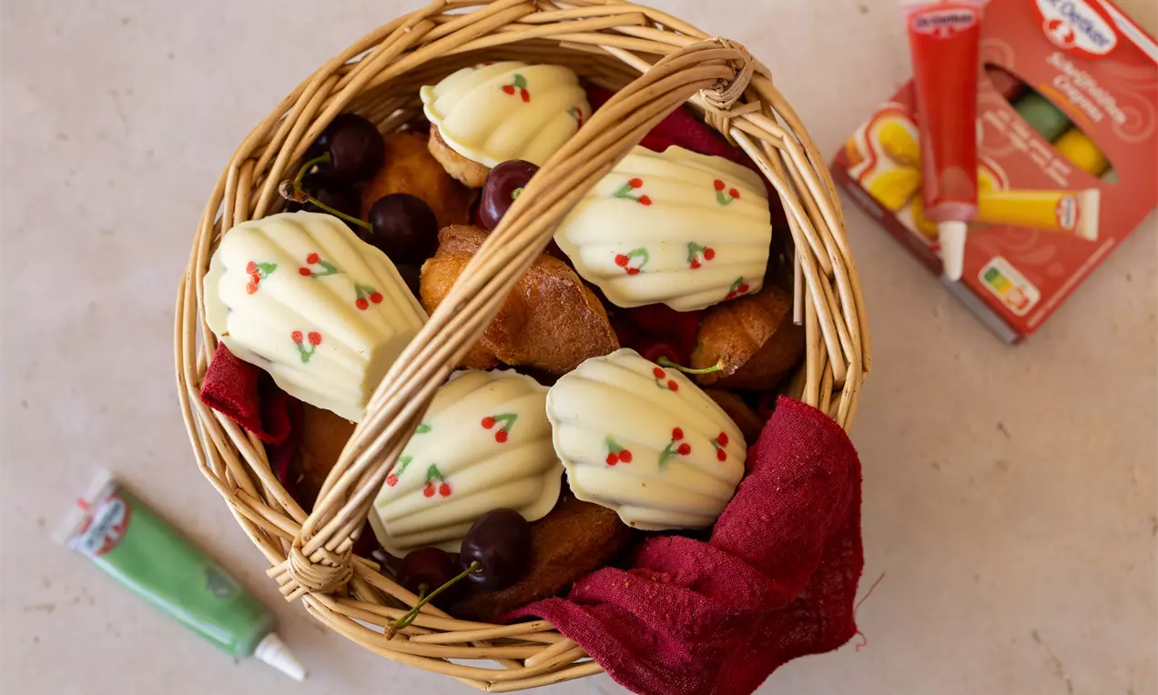 Madeleines met witte chocolade en rode en groene decoratie, licht gebogen en gegarneerd, in een rieten mand met rode doek en kersen