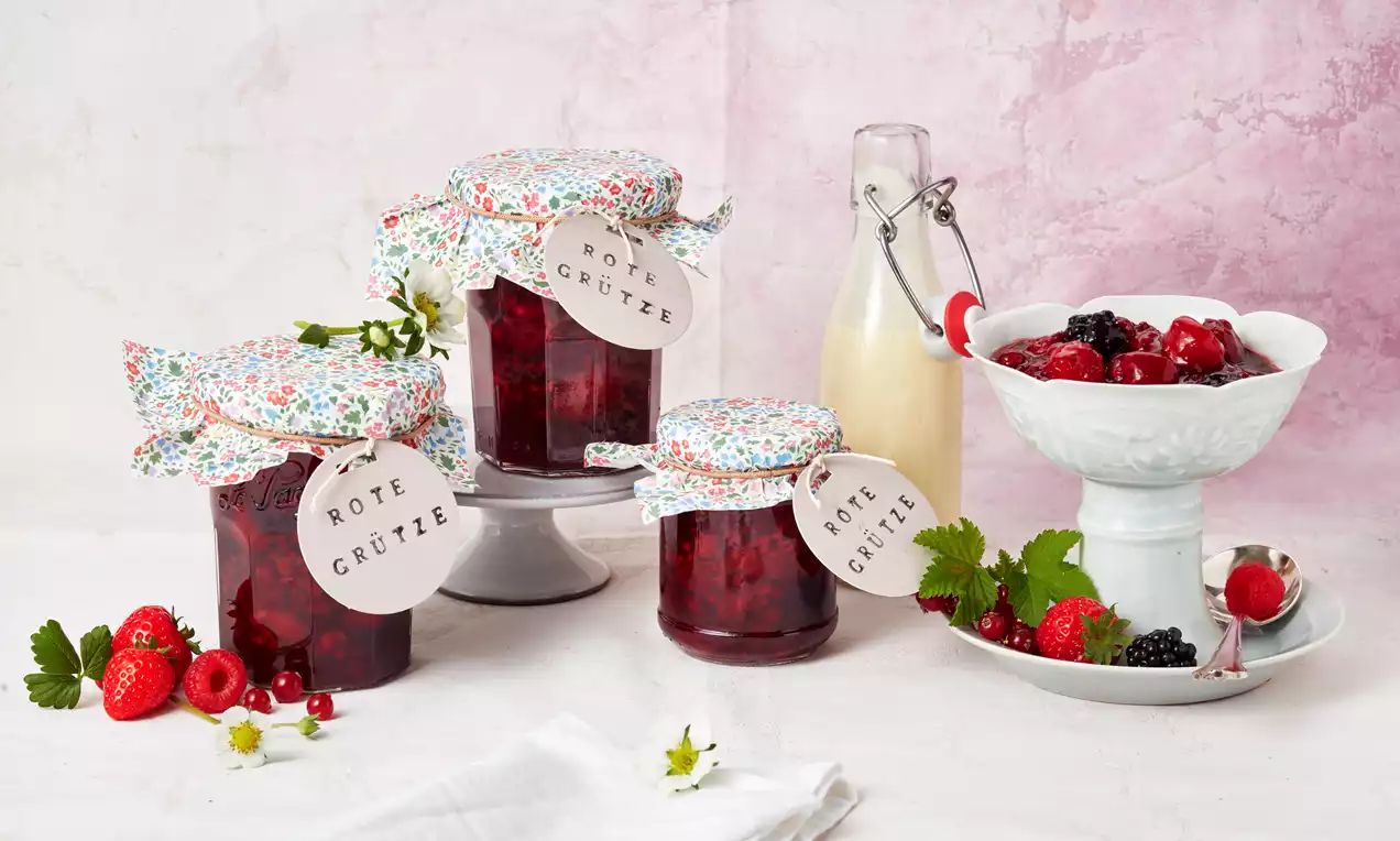 Eingekochte rote Grütze in gläsernen Marmeladengläsern mit bunter Haube, arrangiert mit frischen roten Beeren und weißer Milchflasche