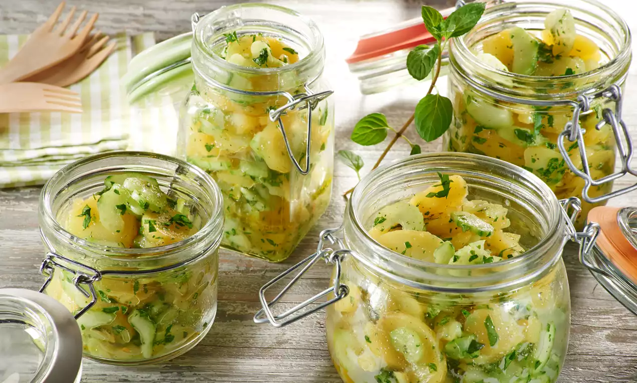 Goldgelber Kartoffelsalat mit grüner Petersilie, Gurkenscheiben und Zwiebelringen in vier offenen Einmachgläsern mit Bügelverschluss drapiert